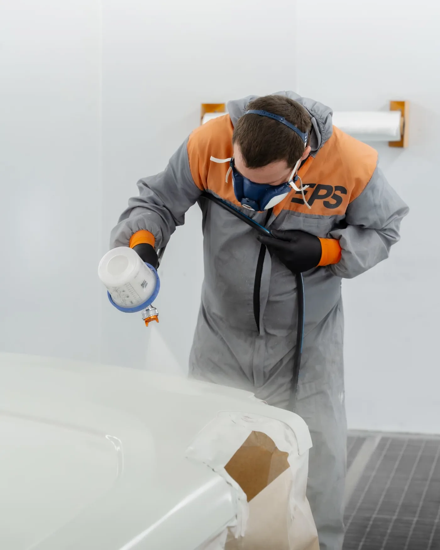 Peintre en carrosserie, masque et combinaison, appliquant la peinture sur un élément de véhicule en cabine