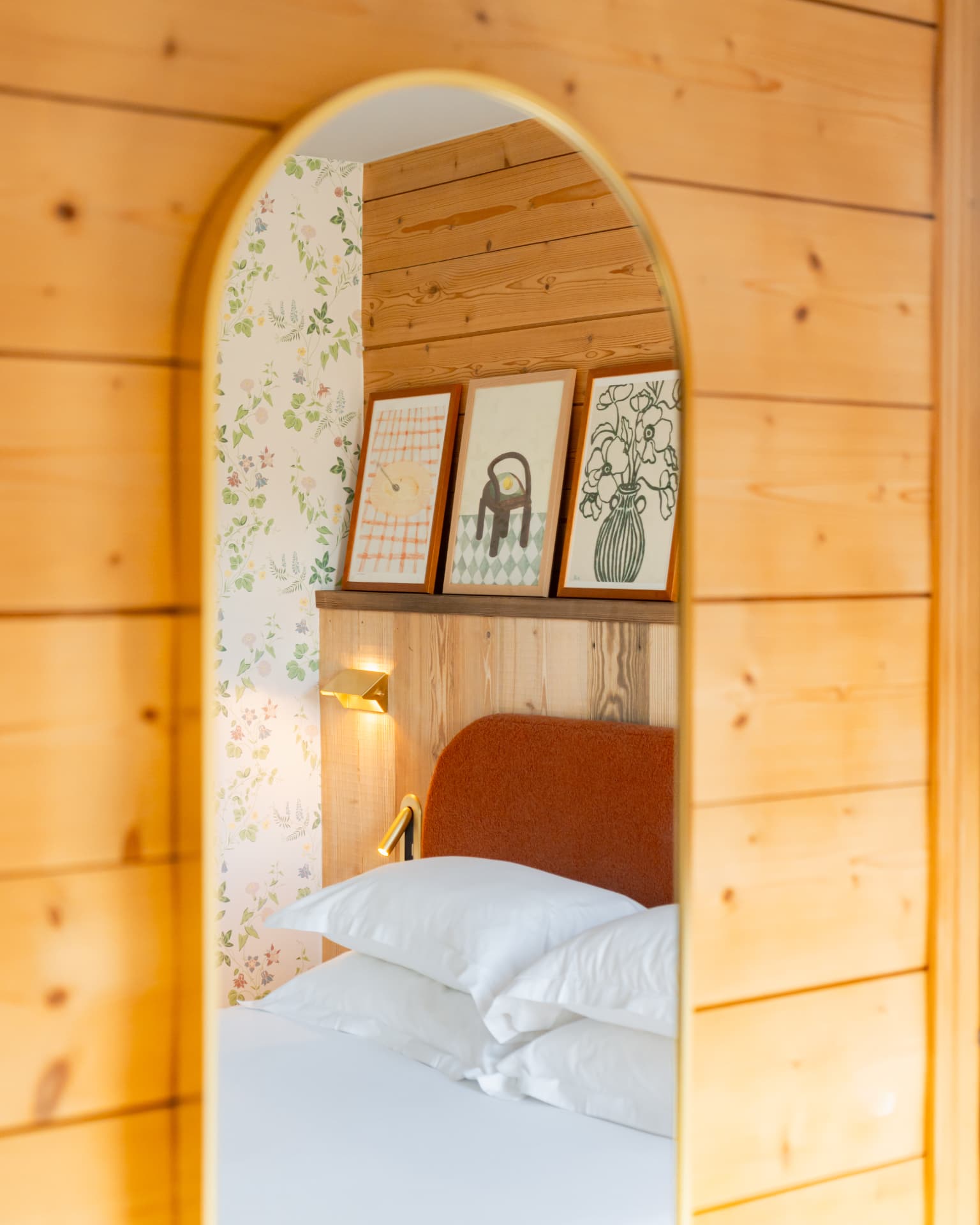Alcôve en bois clair ouvrant sur un lit aux draps blancs, papier peint fleuri et cadres posés sur une étagère