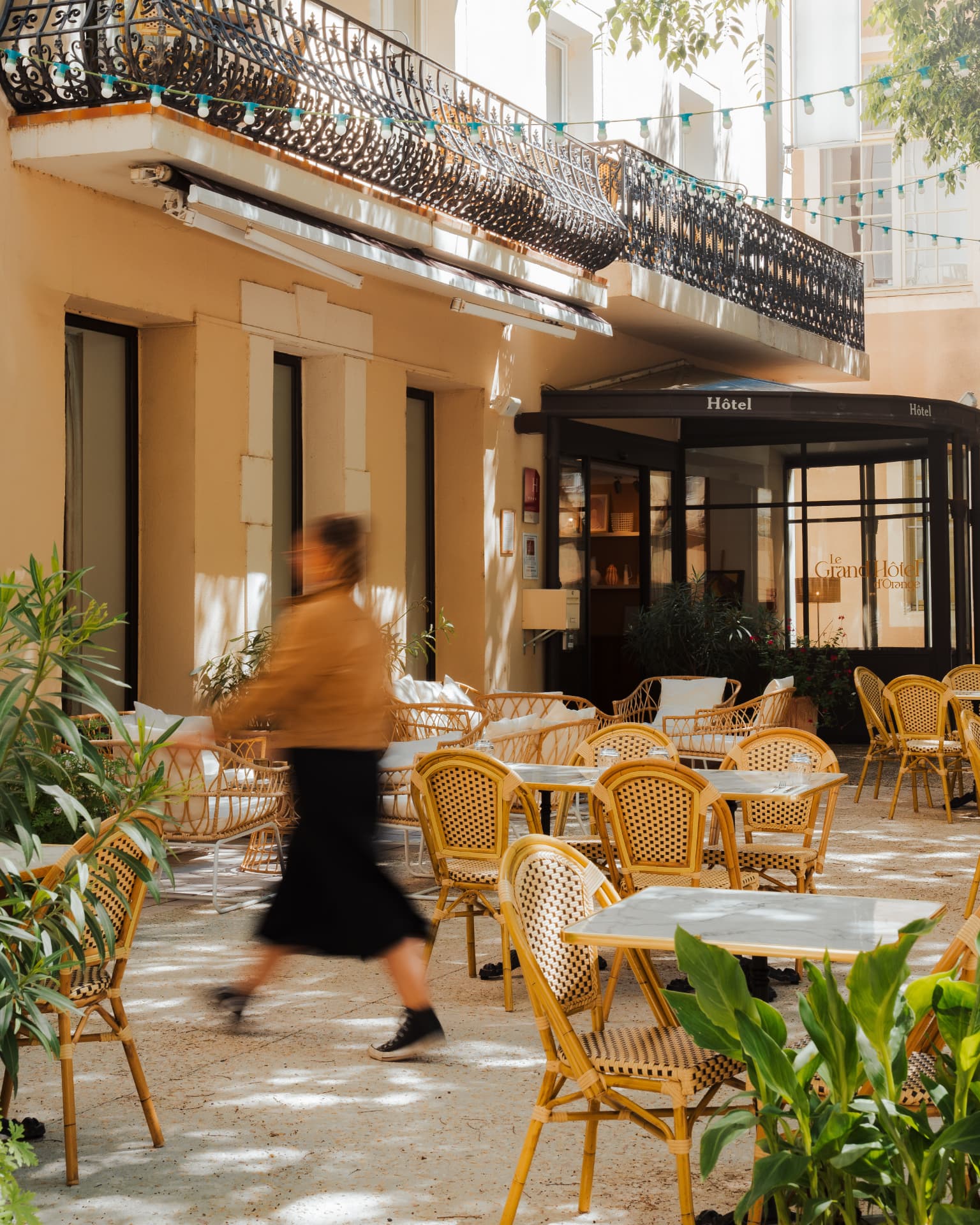 Patio ombragé d'un hôtel avec tables et chaises en rotin, membre de l'équipe traversant la terrasse devant l'entrée