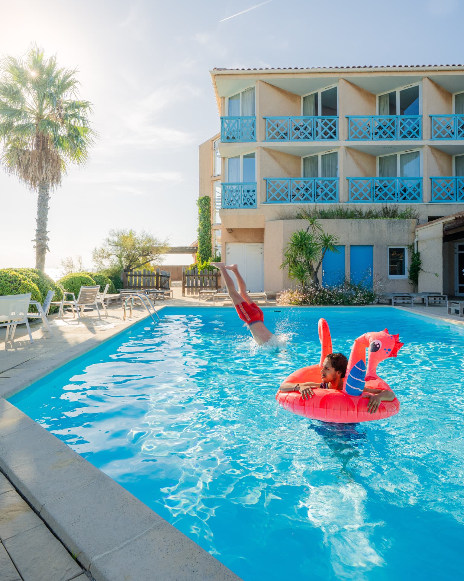 Piscine extérieure d'un hôtel en bord de mer, palmier et balcons bleus, deux voyageurs se baignent au soleil