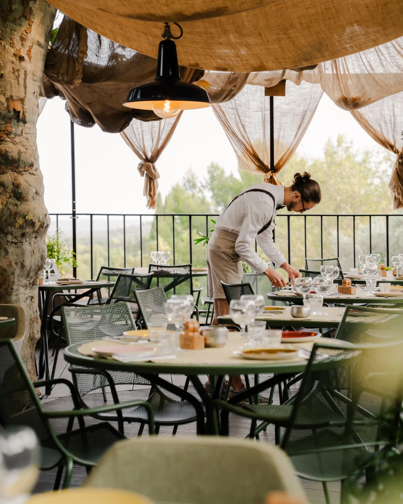 Terrasse de restaurant dressée sous une voile d'ombrage, un membre de l'équipe finalise la mise en place avant le service