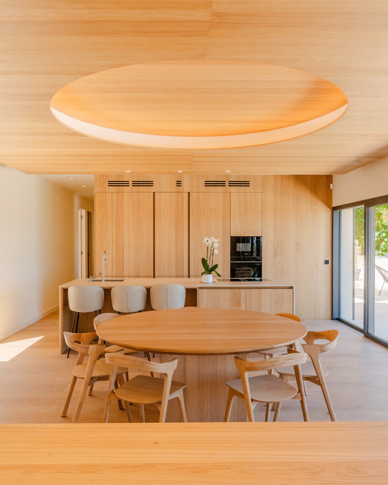 Grande pièce de vie contemporaine habillée de bois clair, table ronde et îlot de cuisine ouverts sur une terrasse