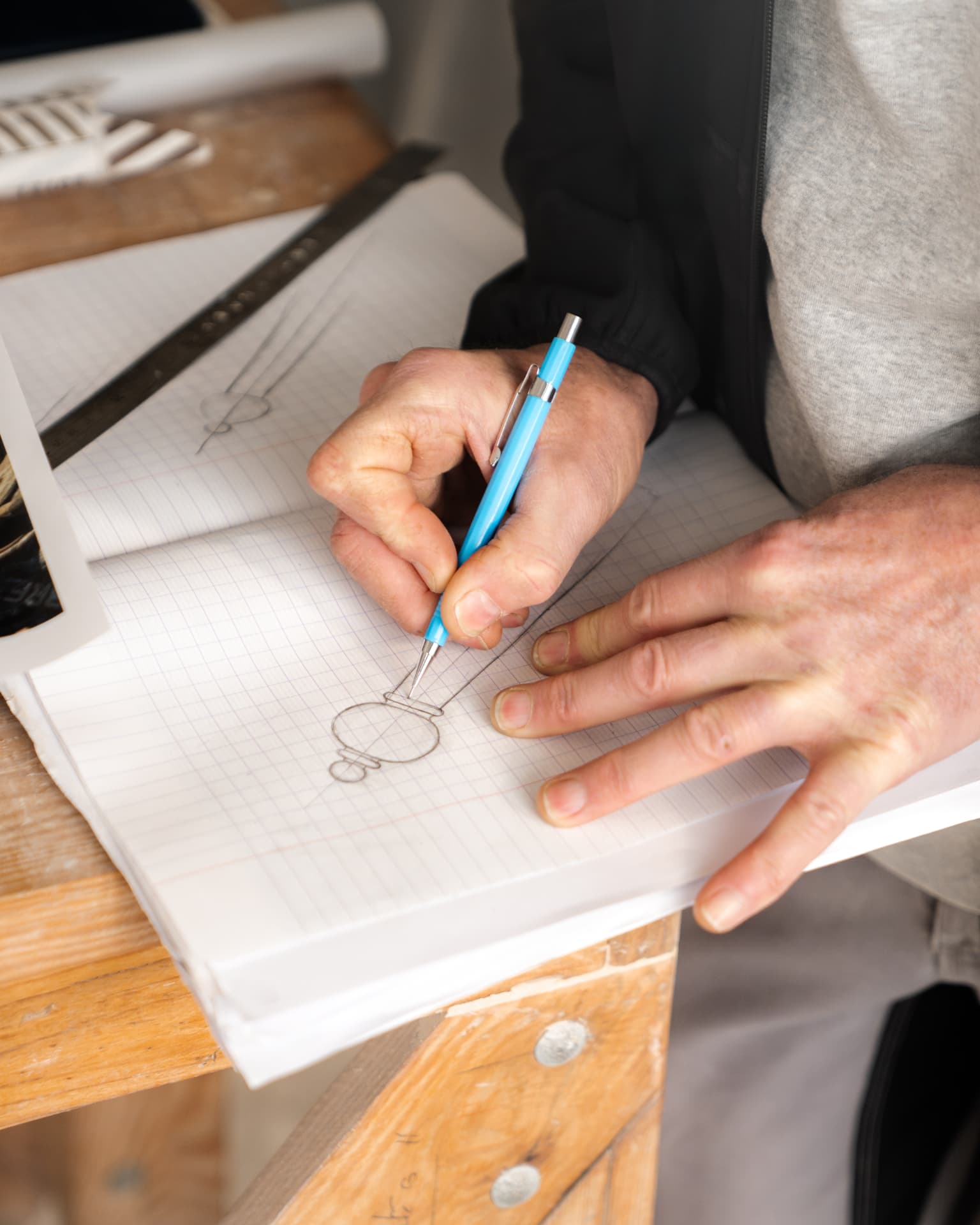 Mains d'un artisan traçant au crayon un dessin technique sur du papier millimétré posé sur un établi en bois