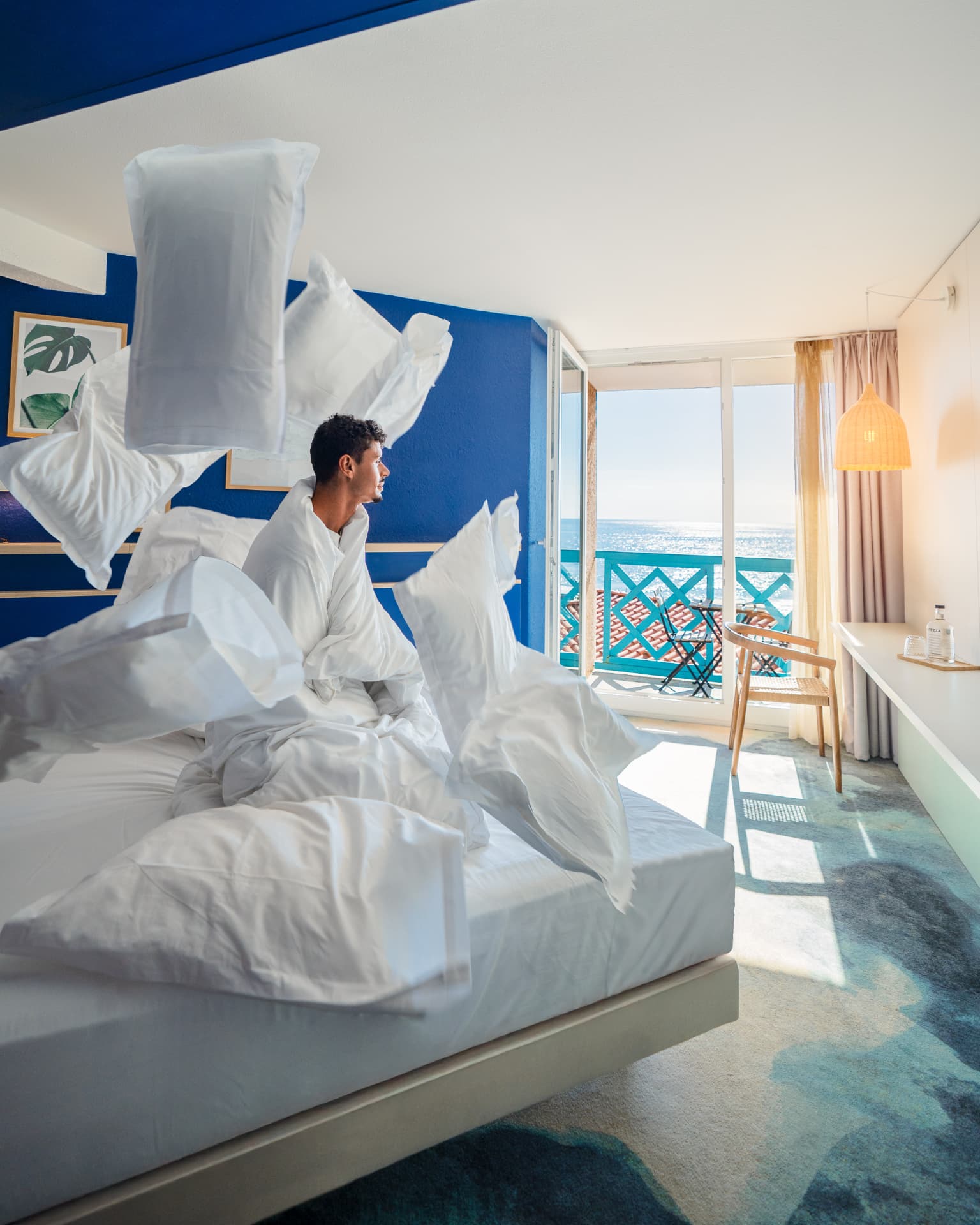 Chambre d'hôtel aux murs bleus ouverte sur un balcon avec vue sur la mer, oreillers blancs en suspension au-dessus du lit