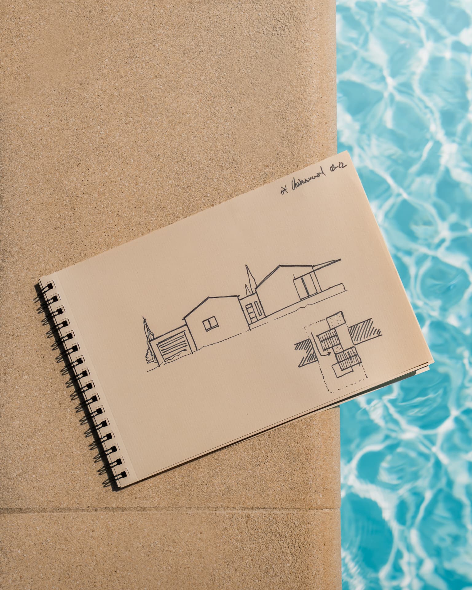 Carnet de croquis ouvert sur un dessin de façade et un plan de masse, posé au bord d'une piscine