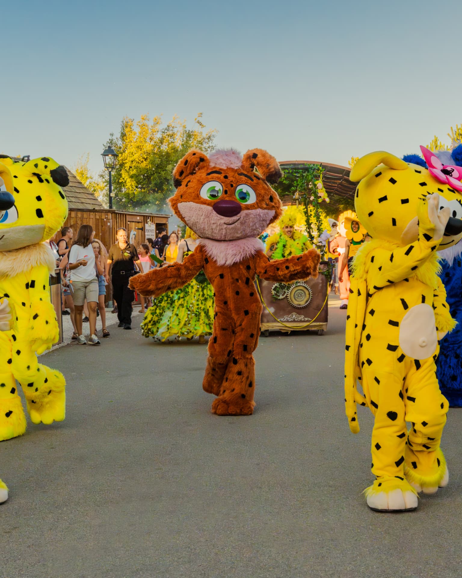 Parade de mascottes costumées avançant dans une allée du parc en fin de journ ée, char fleuri en arrière-plan