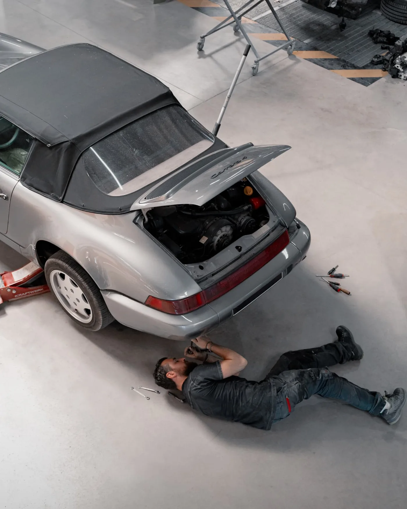 Mécanicien allongé sous l'arrière d'un cabriolet de collection, capot moteur ouvert, dans un atelier