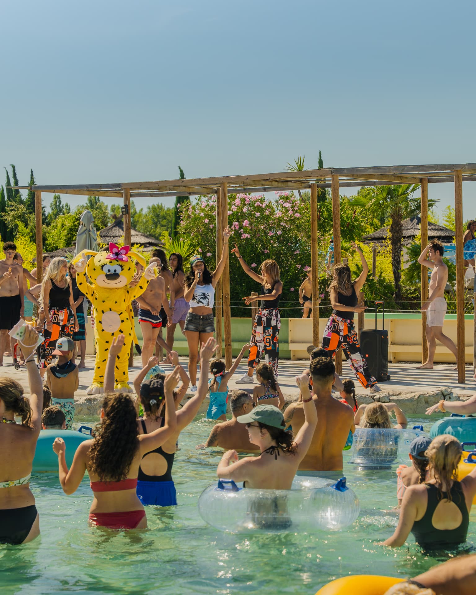 Animation musicale au bord d'une piscine d'un parc de loisirs, mascotte et animateurs face aux visiteurs qui lèvent les bras dans l'eau