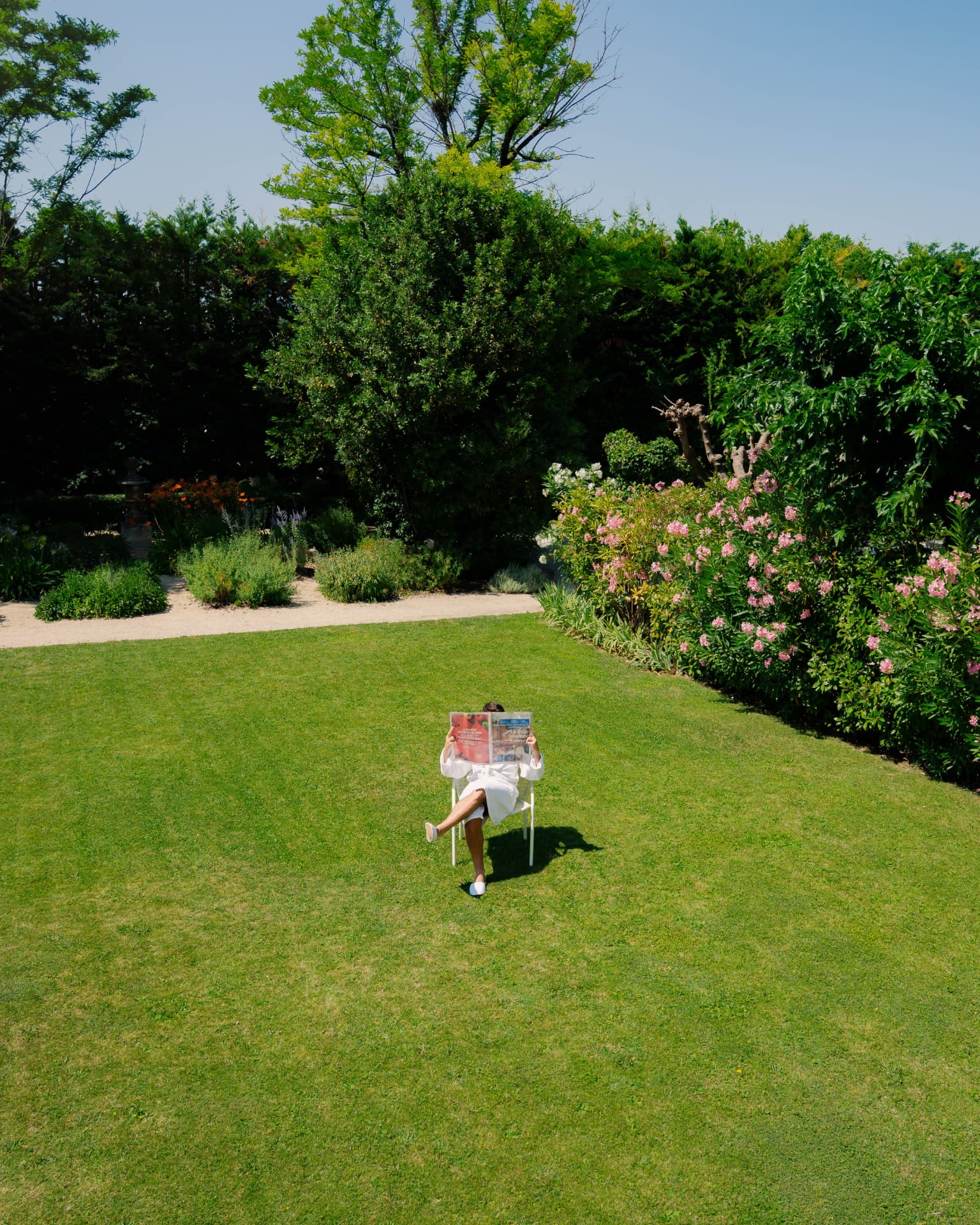 Grand jardin clos et fleuri, une personne lit un journal installée sur une chaise au milieu de la pelouse