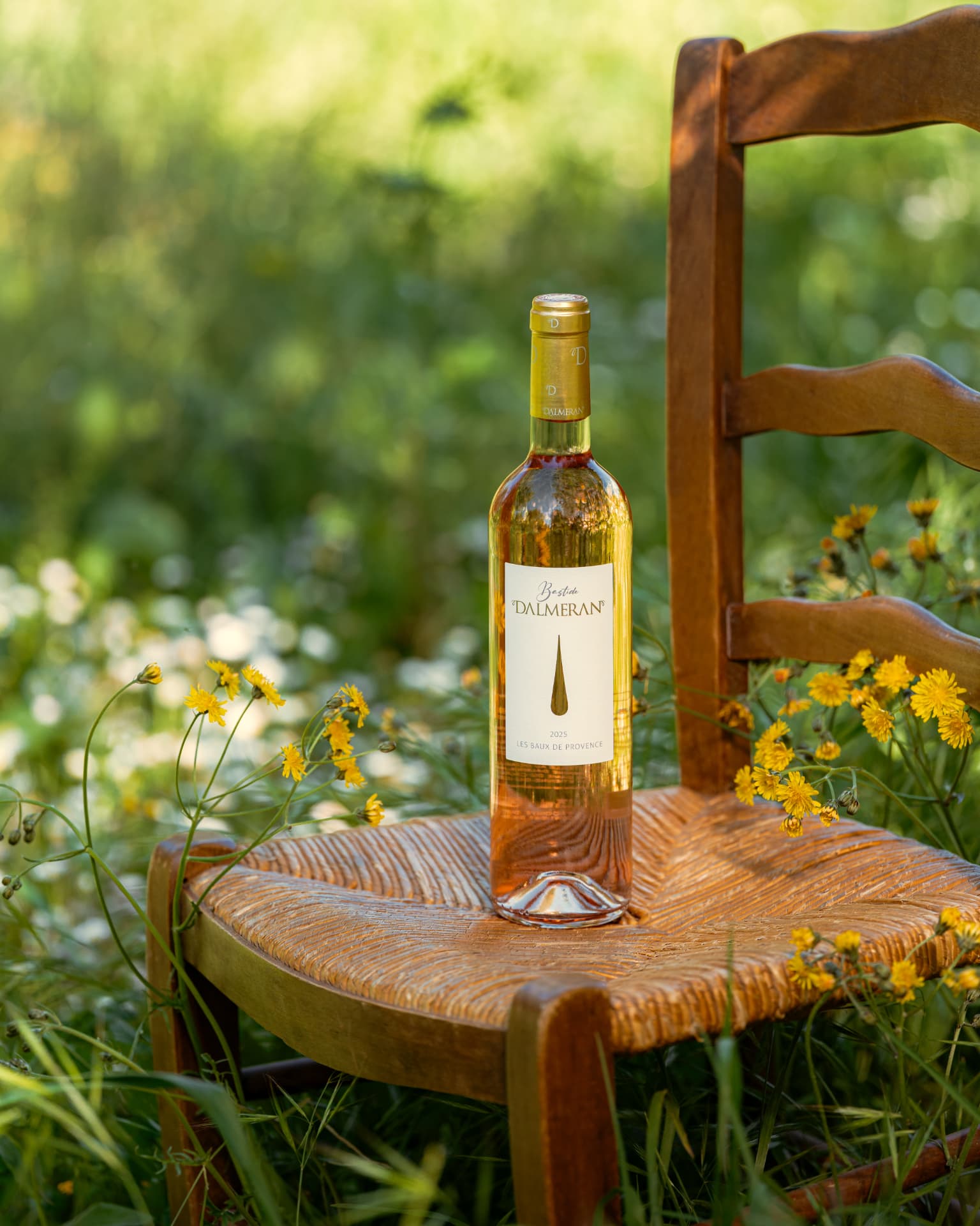 Bouteille de vin rosé posée sur une chaise paillée, au milieu de fleurs sauvages jaunes en pleine lumière